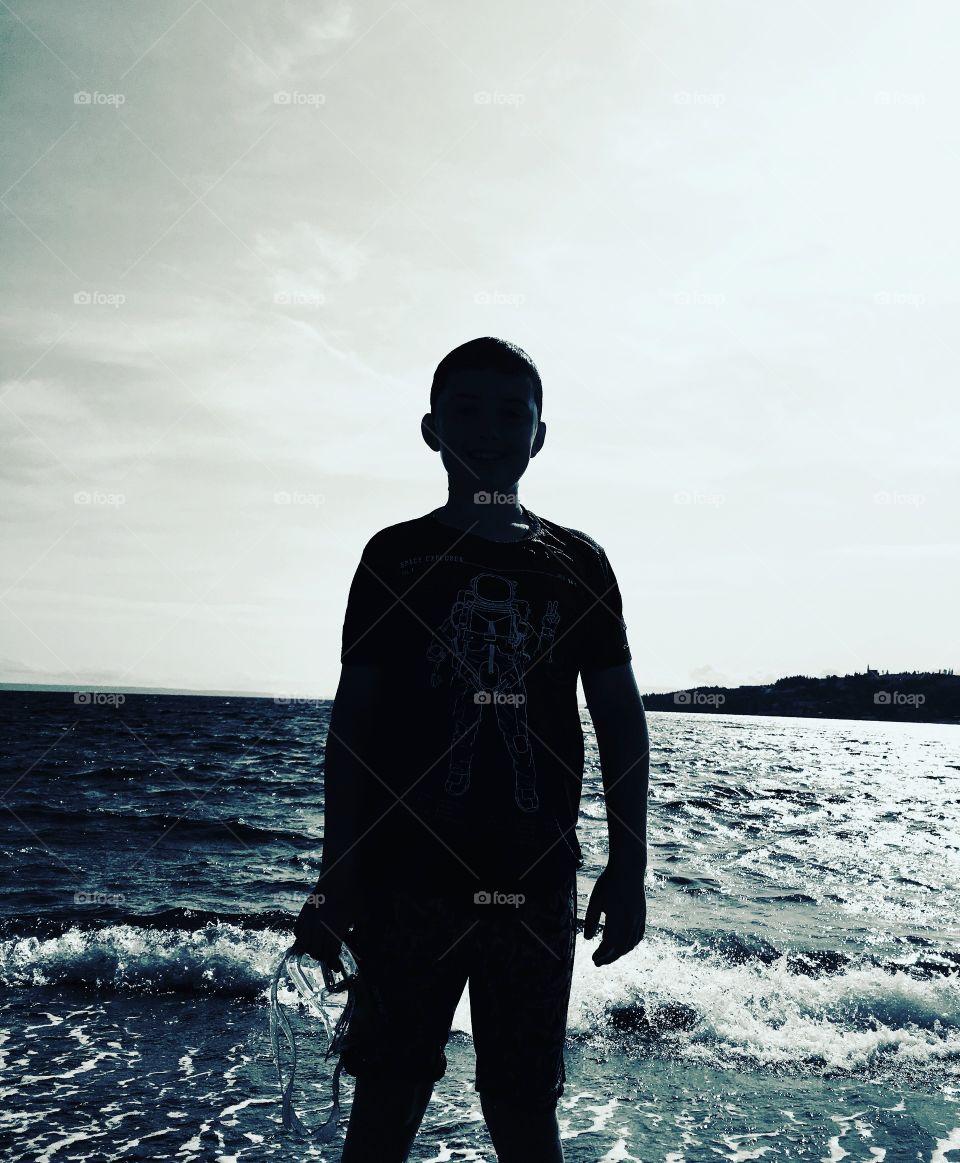 Silhouette of a Boy coming out of the water at the beach holding goggles. Black and white. 