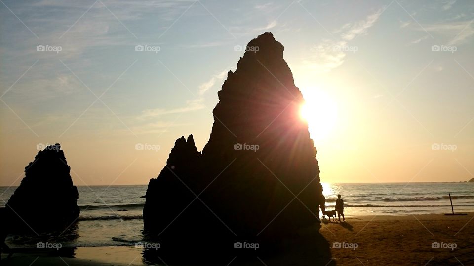 Sun rays meeting tall rock in front of ocean