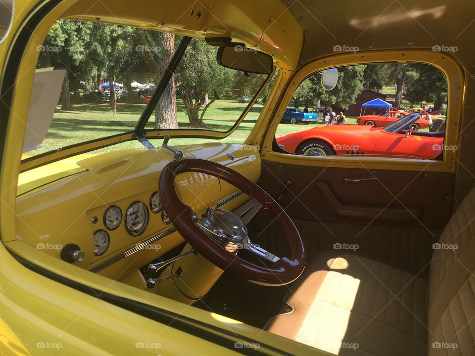 Dashboard and interior of a yellow classic car. Another classic car and a few people are visible in the background. 