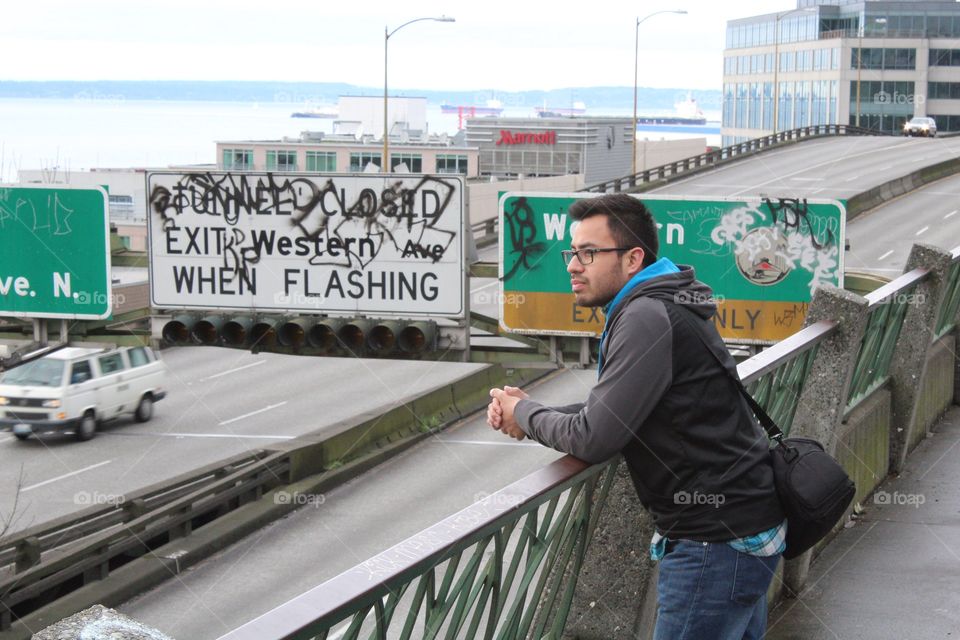 Young Hispanic man pensively stares across an urban interstate freeway in Seattle, Washington with graffitied signs in the background