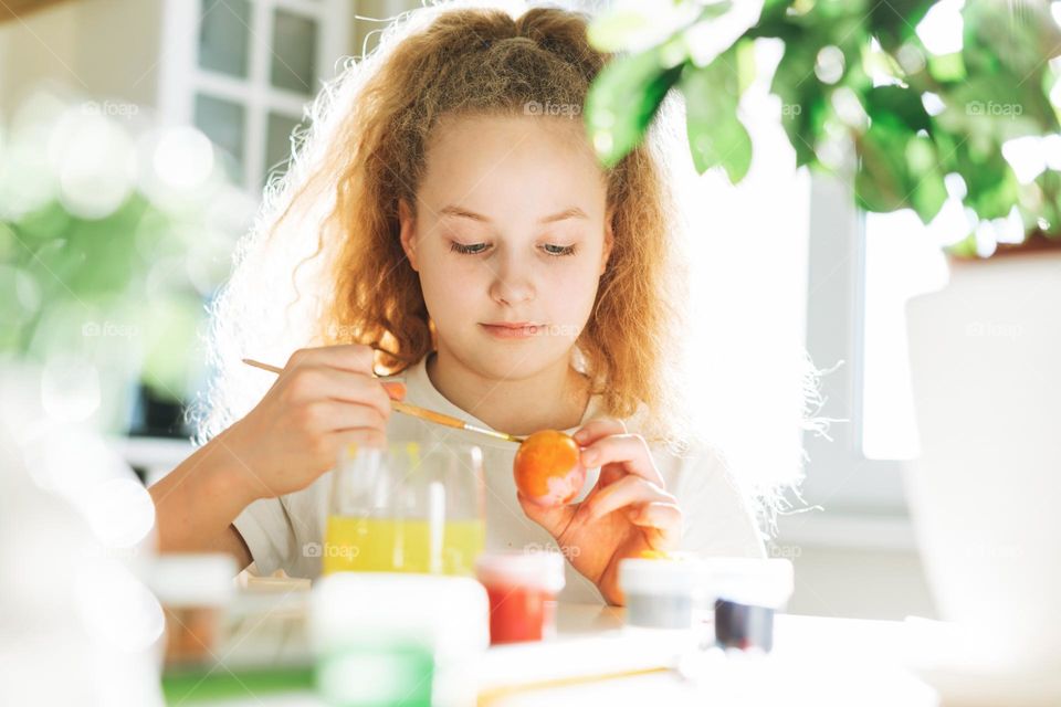 Cute tween girl with long curly hair painting eggs on kitchen at home on spring sunny day, Happy Easter