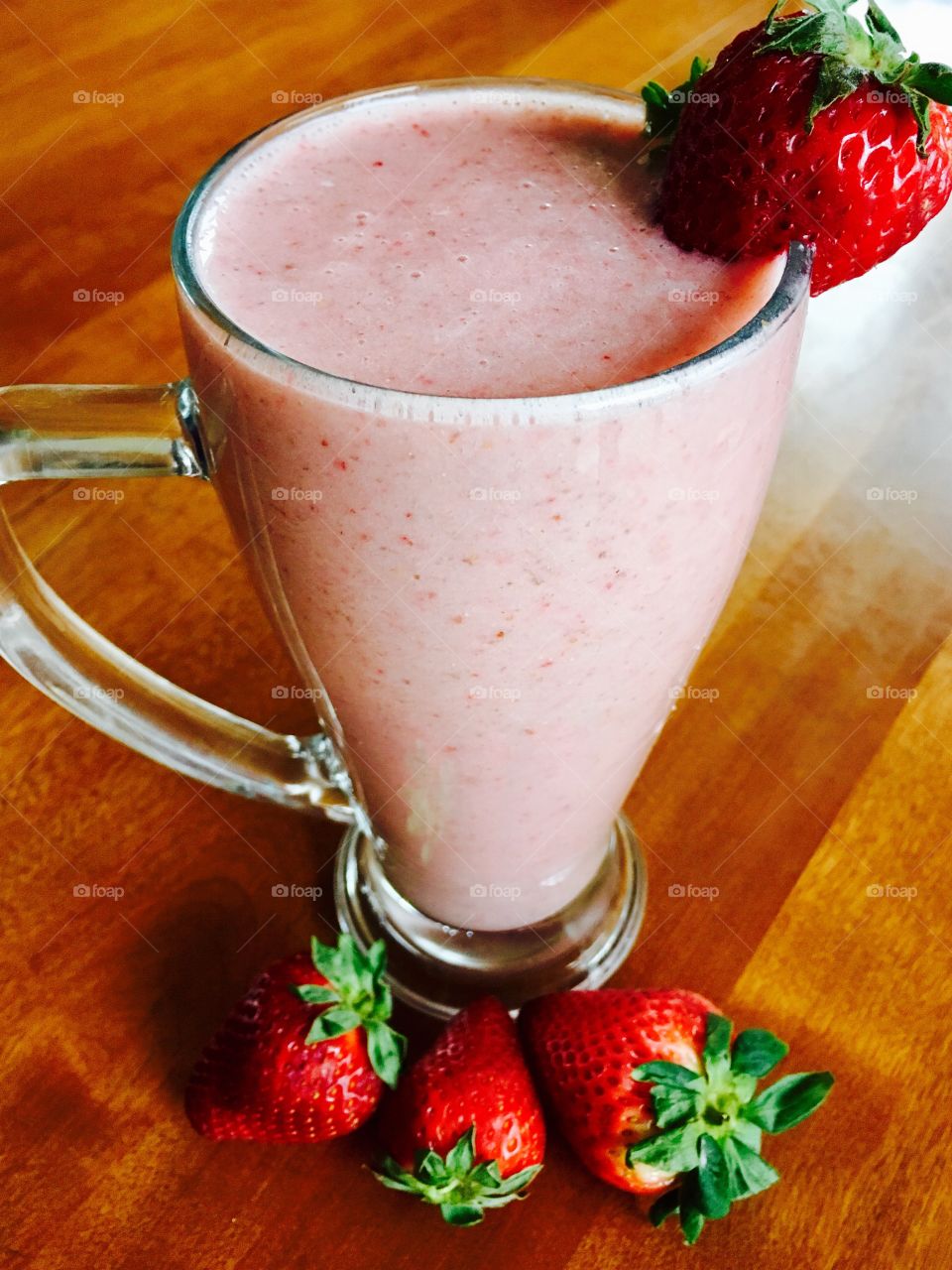 Fresh strawberry and banana smoothie in a tall glass. Garnished with a strawberry and surrounded by more berries on a wooden table. 