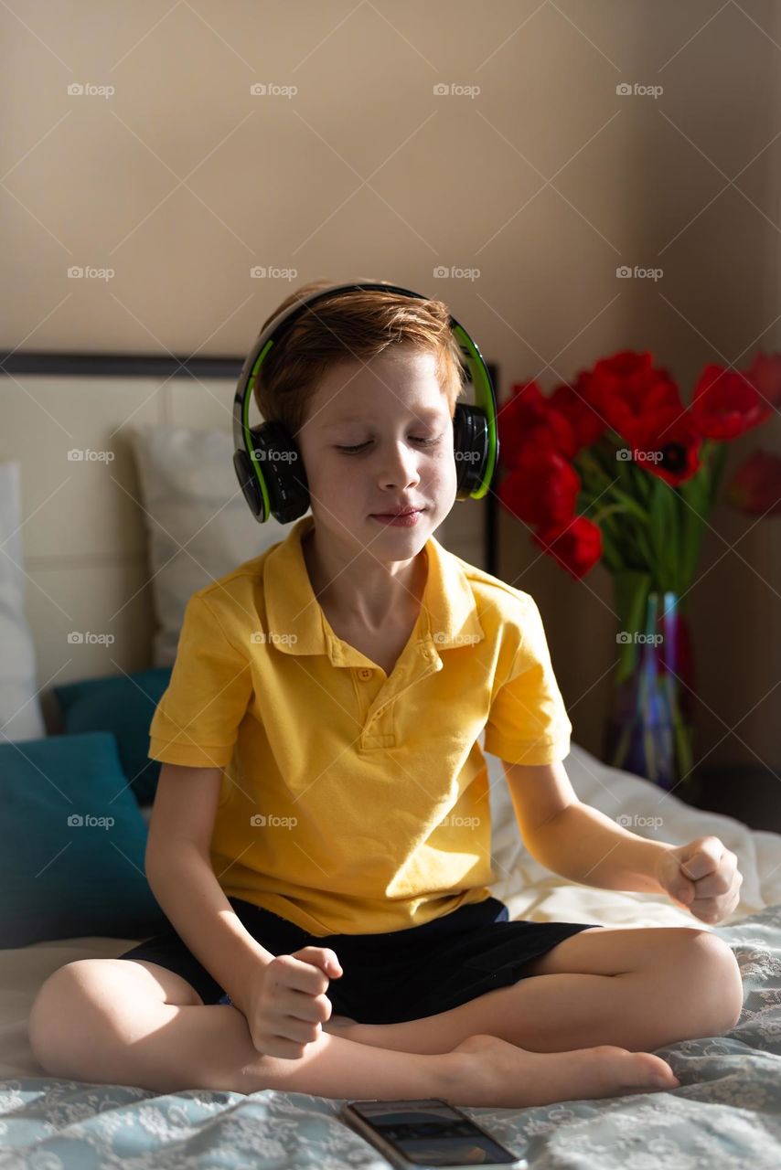 red-haired cute child boy sitting at home on the bed in headphones listening to music