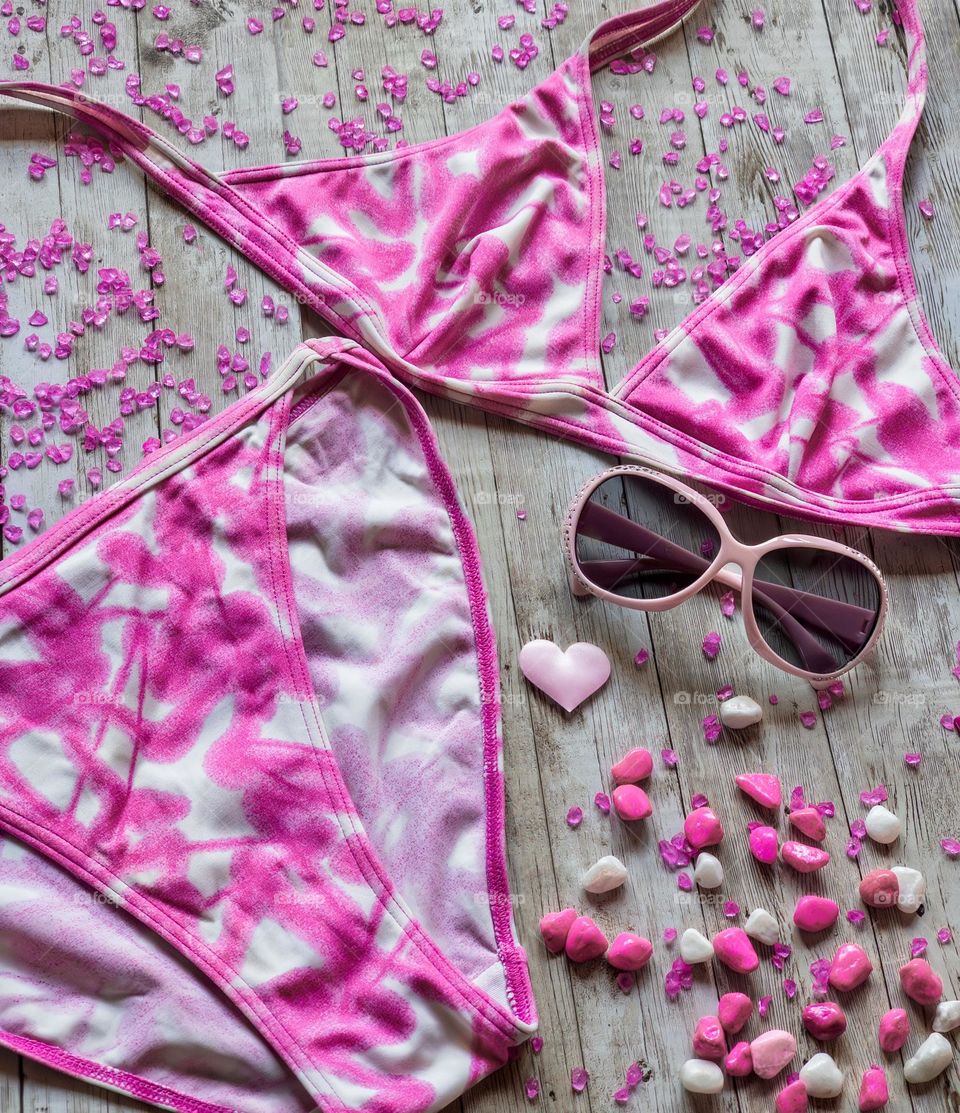 Shocking pink & white bikini, with sunglasses surrounded by pink pebbles & gravel on wood background 