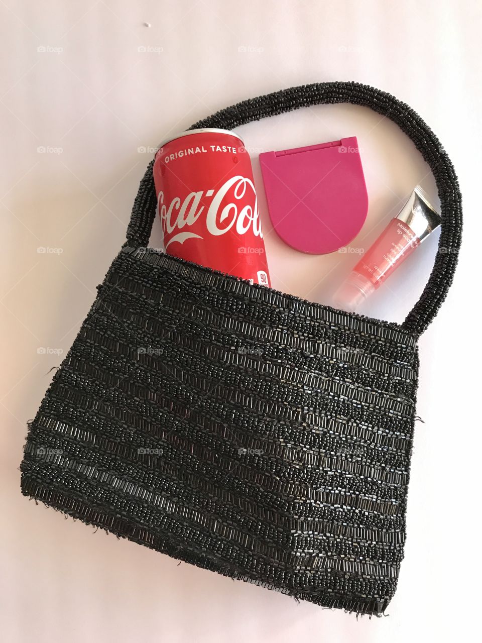 A can of Coca Cola with a pink compact and pink lip glass with a black purse on a white background flat lay