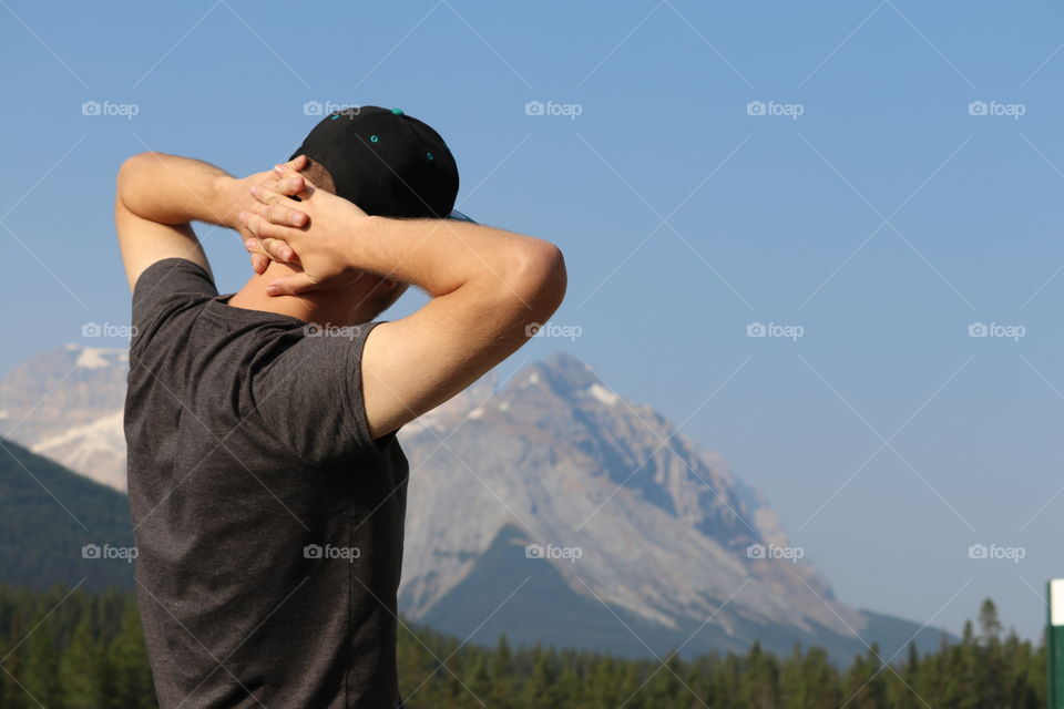 Young man in his twenties with arms clasped behind his head viewing Canada's beautiful snow-capped Rocky Mountains 