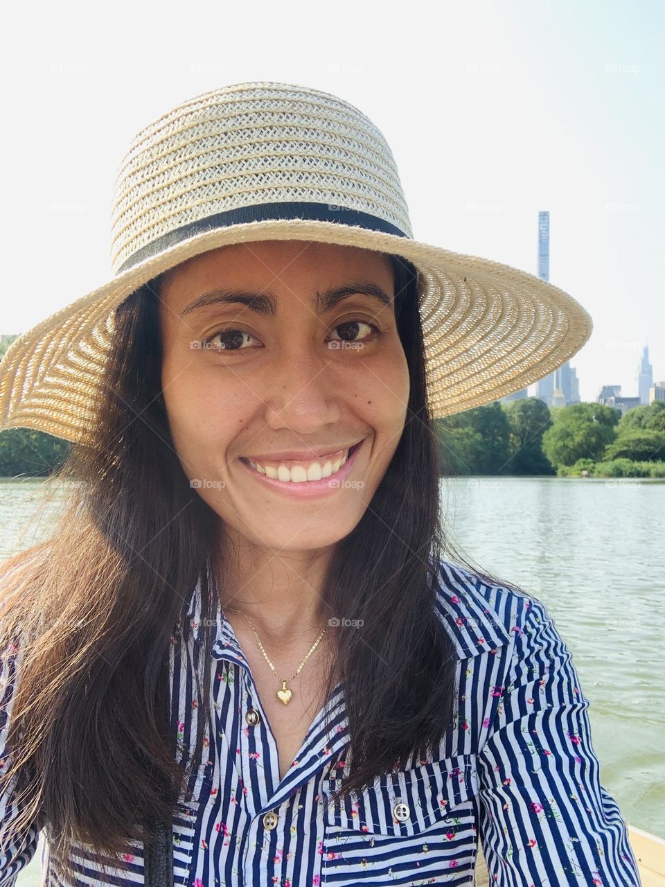 A woman smiling wearing a hat, stripe shirt,sitting on the boat against the sky.