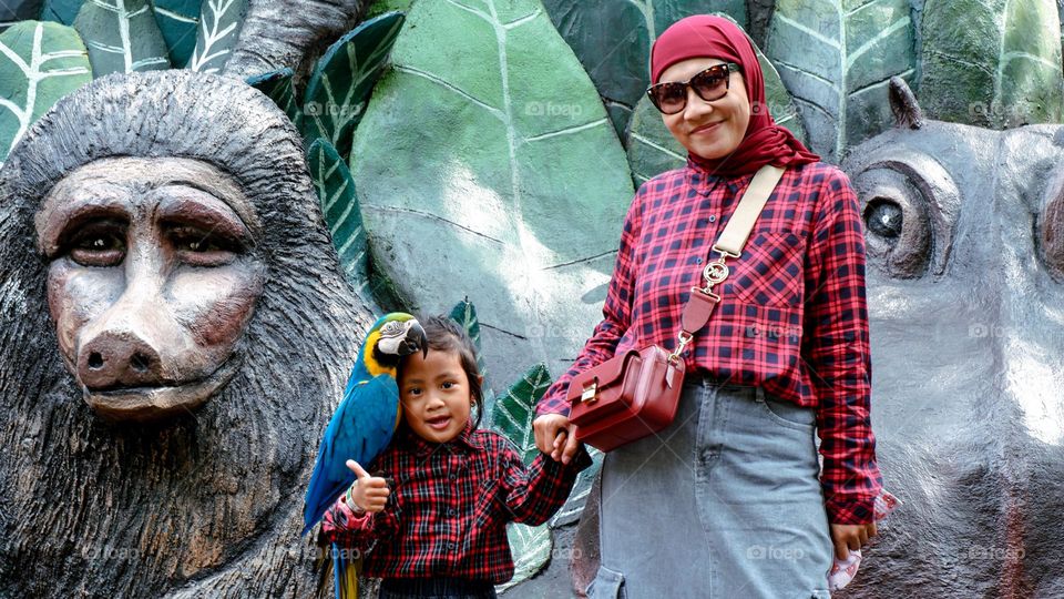 Batu, Indonesia - March 31, 2024 : Portrait of a little girl with her mother taking a photo with a parrot on her shoulder at Batu Secret Zoo, vacationing at the zoo with happy expressions.