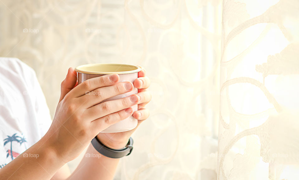 Hands of person hug a cup with beverage near the window. Concept of cozy home.