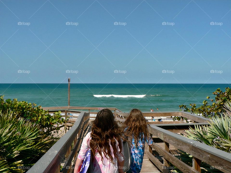 Two young children girls carrying body boards going on the boardwalk walking towards the beach on the Atlantic Ocean wavy beach with blue clear sky.