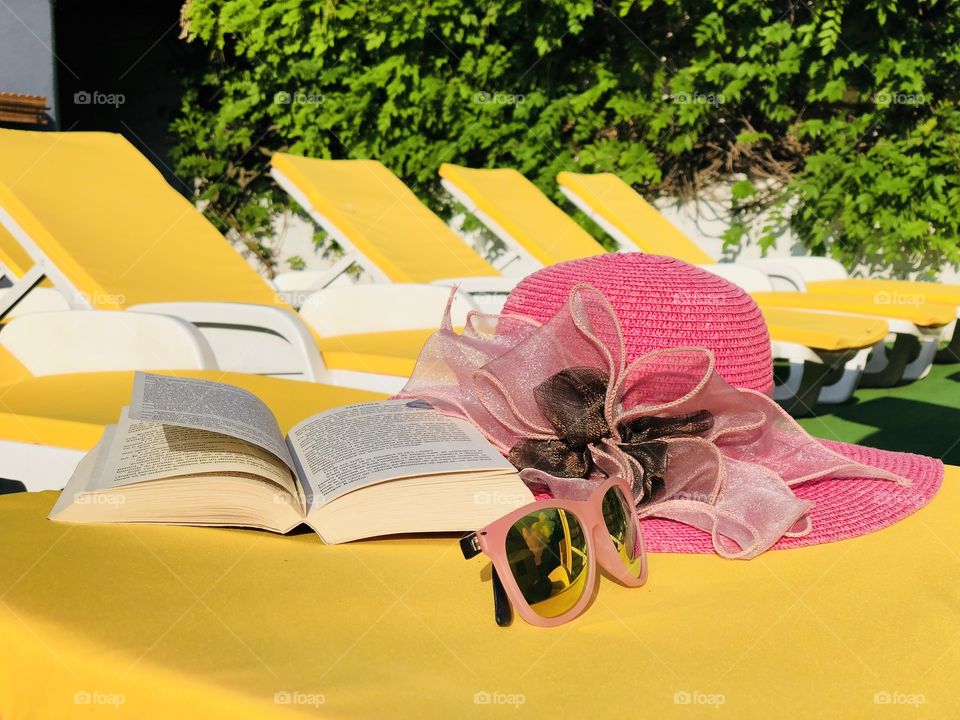 Pink sunglasses, a book and a hat placed on a yellow sunbed with many yellow sunbeds in the background