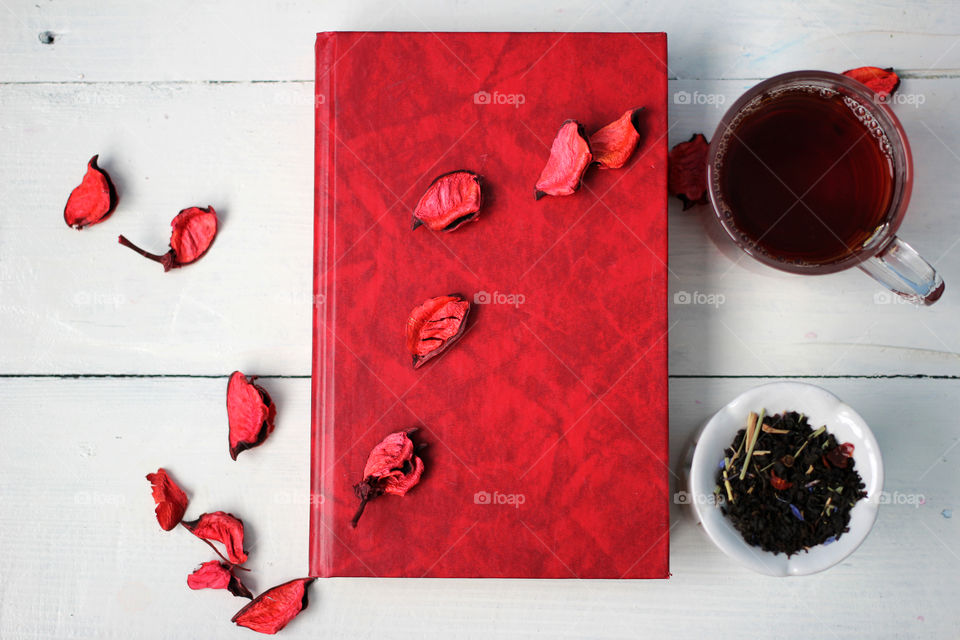 A cup of tea and a book on a white table, which is decorated with rose petals