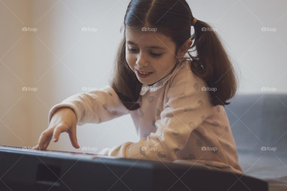 Portrait of a beautiful little caucasian girl with two ponytails on her head playing the electric piano sitting at the sofa in the living room, close-up side view. Music education concept, musical instruments, children playing.