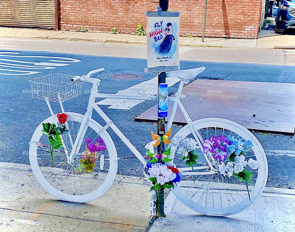 A tragic bicycle accident happened under the „Pulaski Bridge“ at „Borden Ave“ & „11th St“ when a 14 year old was run down by a truck. Since then this bicycle painted white w flowers & his picture has been here at the site. 2022. Hypnotic Productions