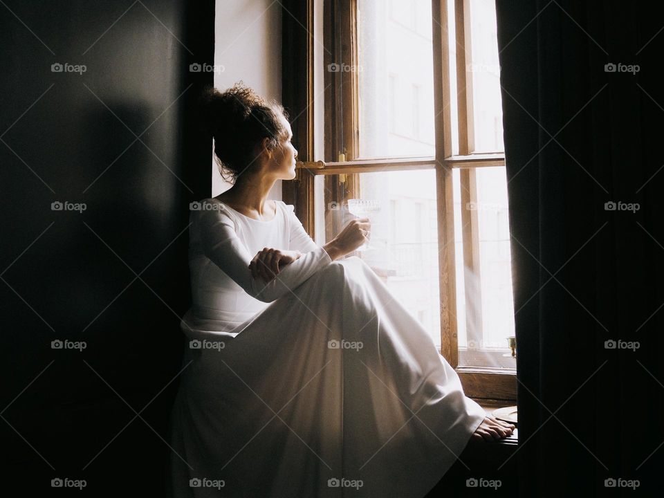 Young beautiful woman with curly hair wearing white dress sits on the window and drinks champagne, portrait of woman, looking at window, lifestyle, at home, relax.