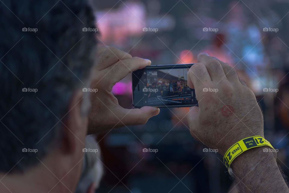 image of a person taking cell phone video in foreground with a blurred outdoor concert setting in the background