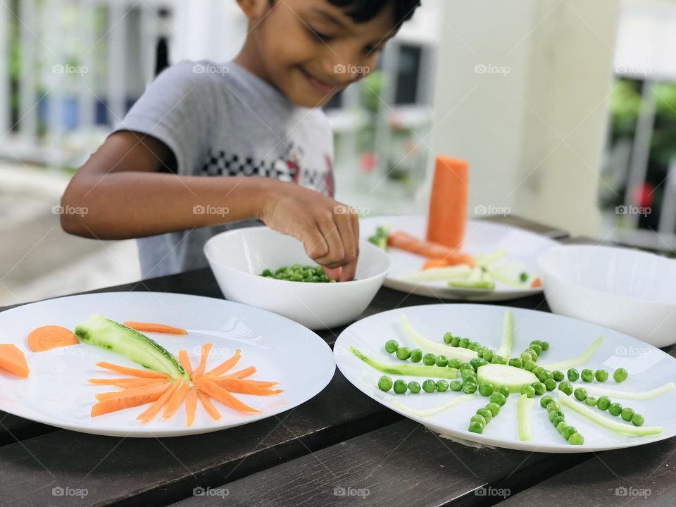 A boy having fun by doing creative craft like coconut tree and sun with vegetables like carrots and cucumbers and Greenpeace.