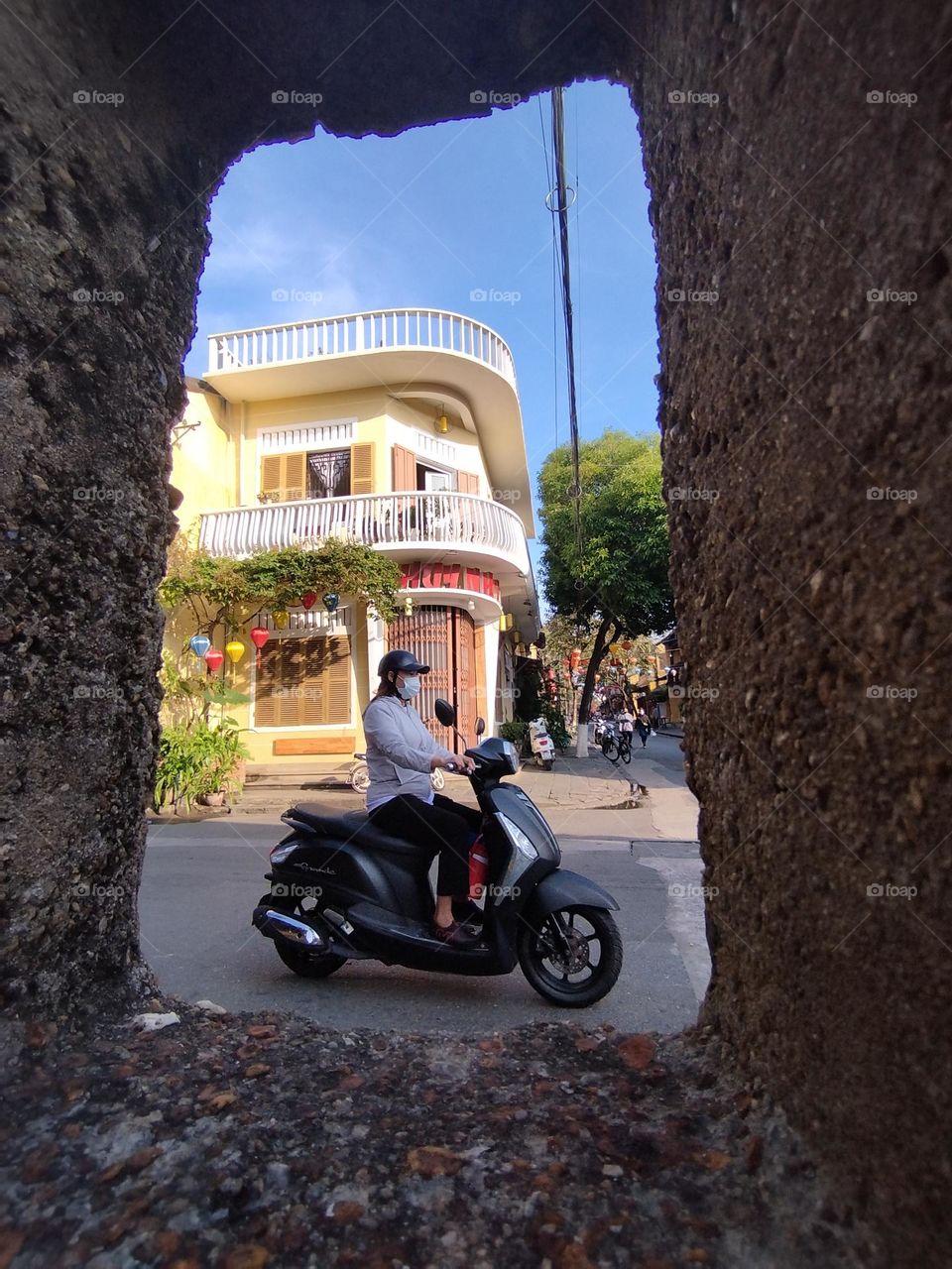 24th January 2025. 08:25am. Location: old town, Hoi an, Vietnam. Person riding a scooter, framed my a concrete post and building in the background.