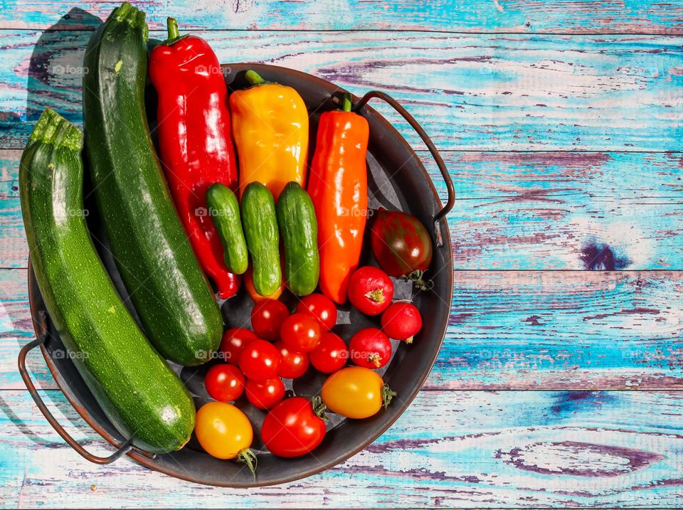 Seasonal vegetables from zucchini, cherry tomatoes, peppers and cucumbers lie in a vintage metal dish on an old shabby deevyan table in blue color, flat lay close-up. The concept of seasonal vegetables, healthy food.