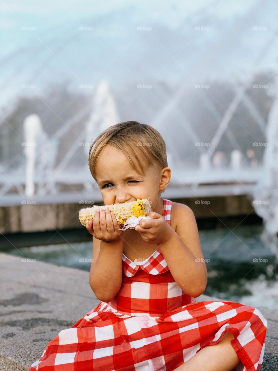 Cute little girl with short blonde hair wearing red dress sitting near fountains and eating corn, portrait of child 