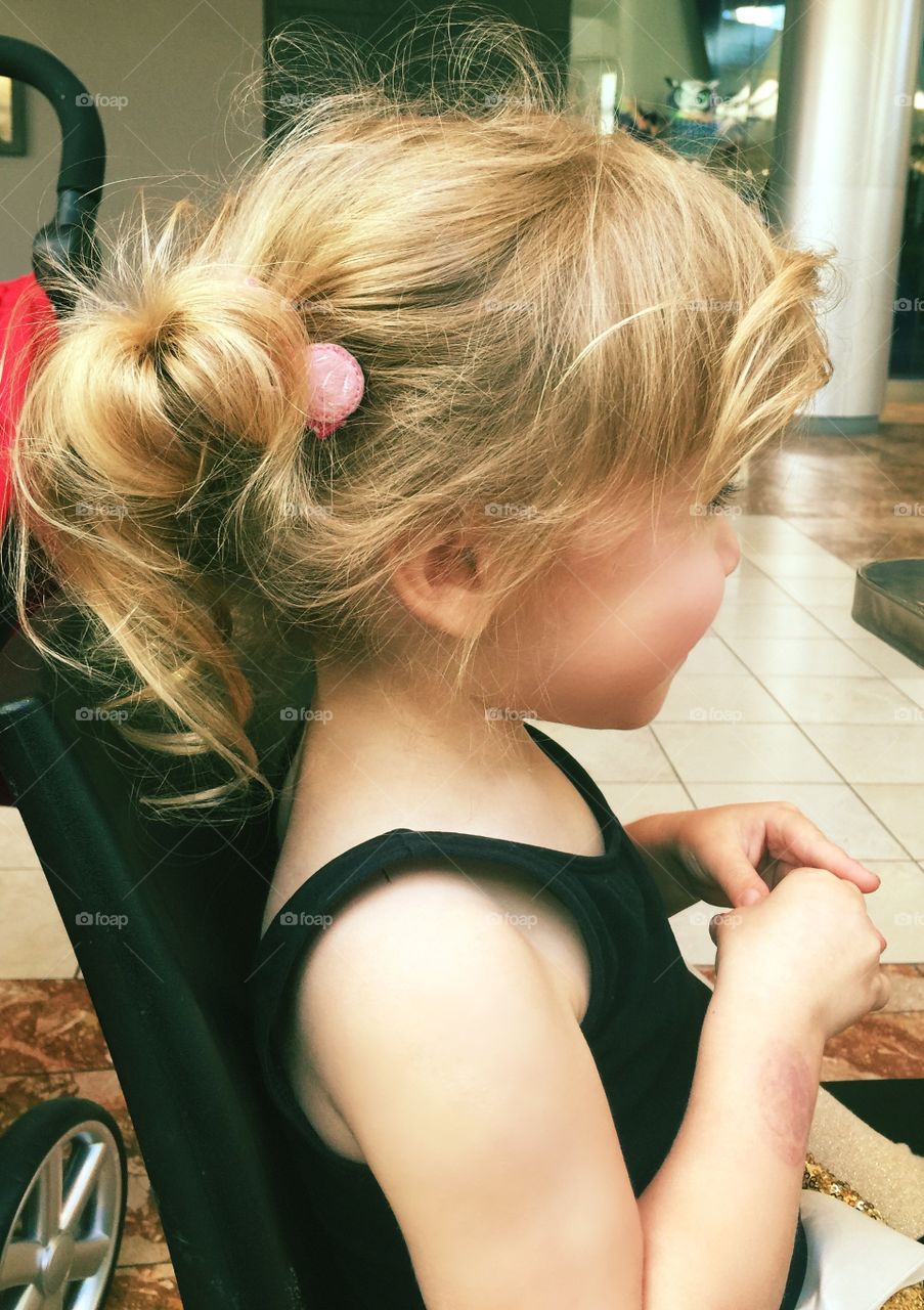 Seated toddler baby girl’s profile with loosely curled blonde pigtails in black tank and pink barrette