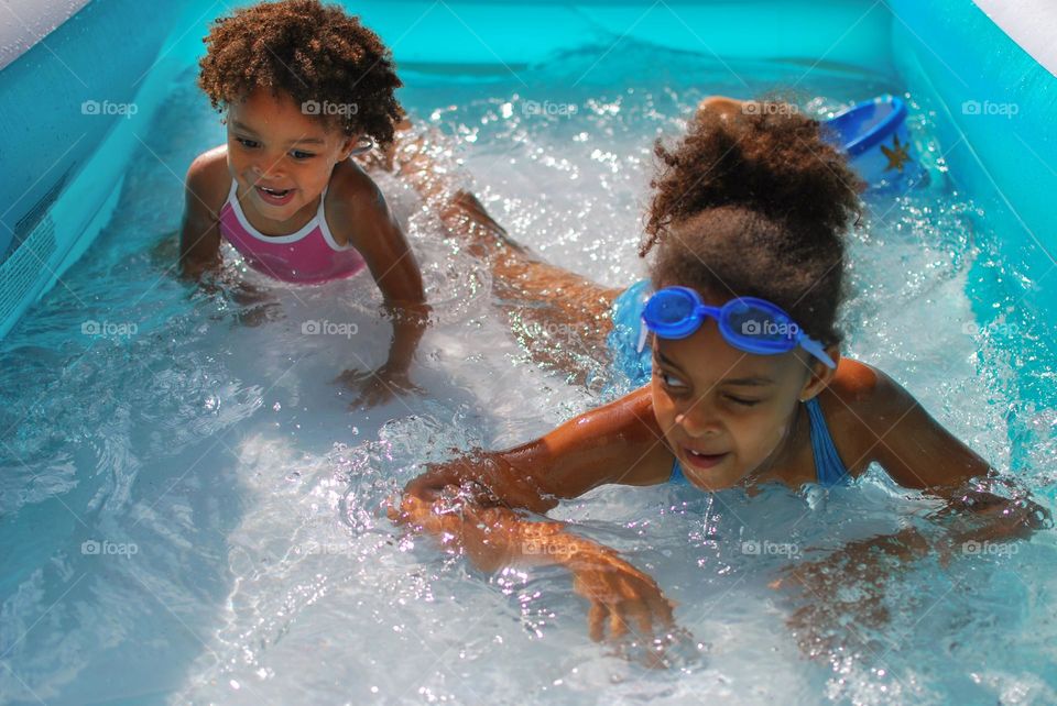 Girl of mixed race enjoying the refreshment of water in a swimming pool on a hot summer day, together with her little sister (family, fun, summer, water, blue, swimming suit, splash, hot, enjoy, play, outdoors)