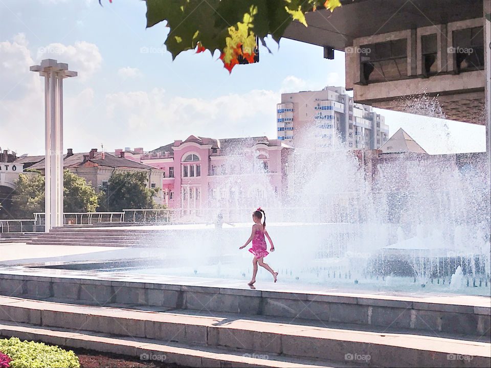 Happy girl running through the splashes of city fountains in summer 