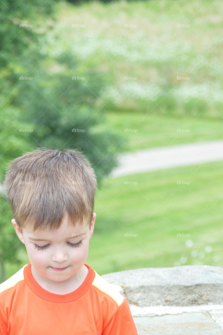 Young boy outdoors looking down in the bottom left of the frame with greenery in the background