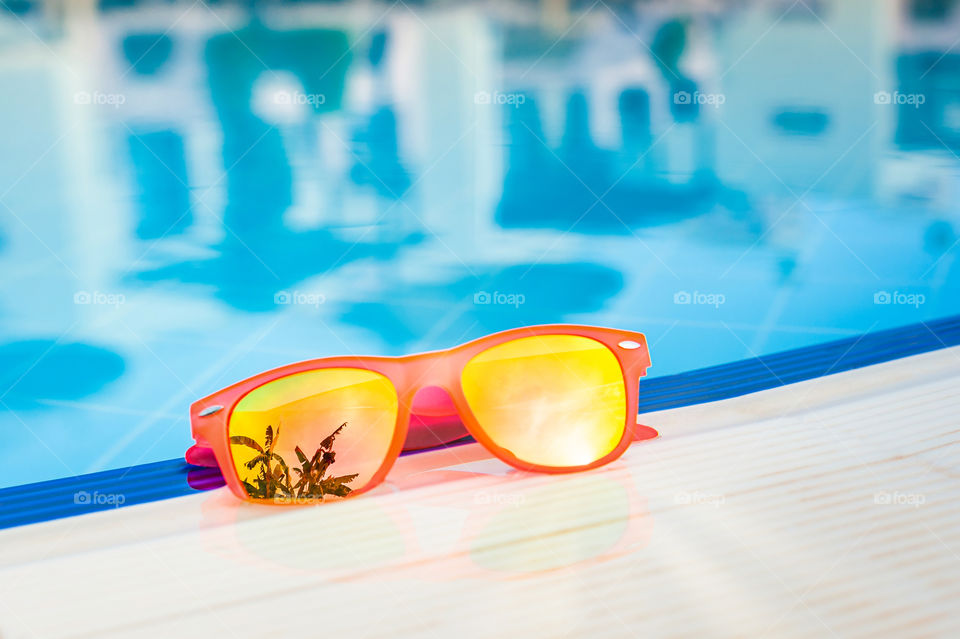 Orange sunglasses reflecting palm-tree and sun left on the edge of the swimming pool.