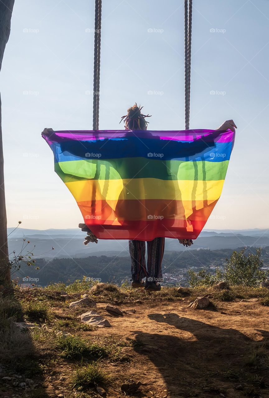 A person on a swing high on a hill holding a rainbow coloured flag