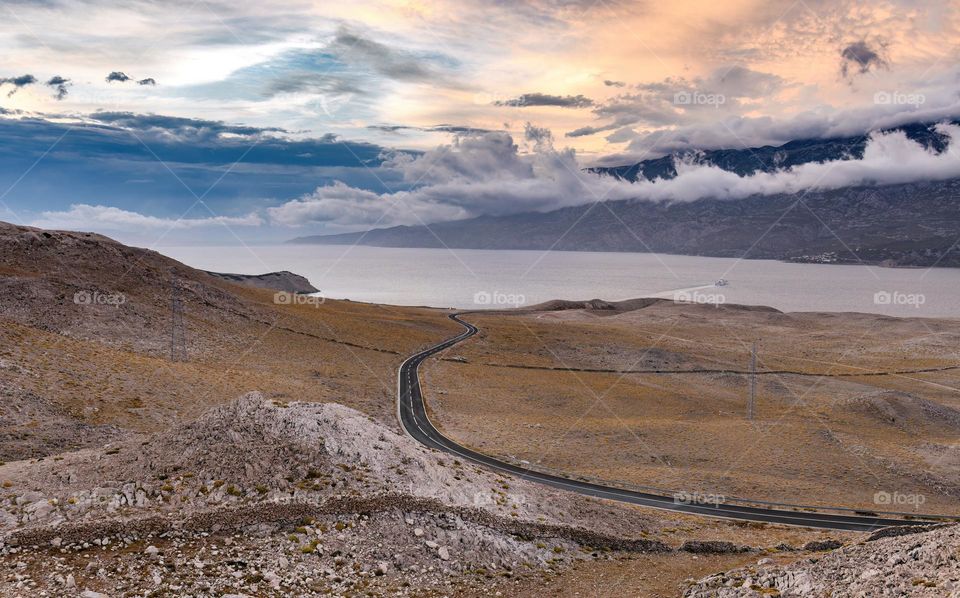 Empty road through arid terrain leading to coast on island of Pag in Croatia. Beautiful clouds above coastline.