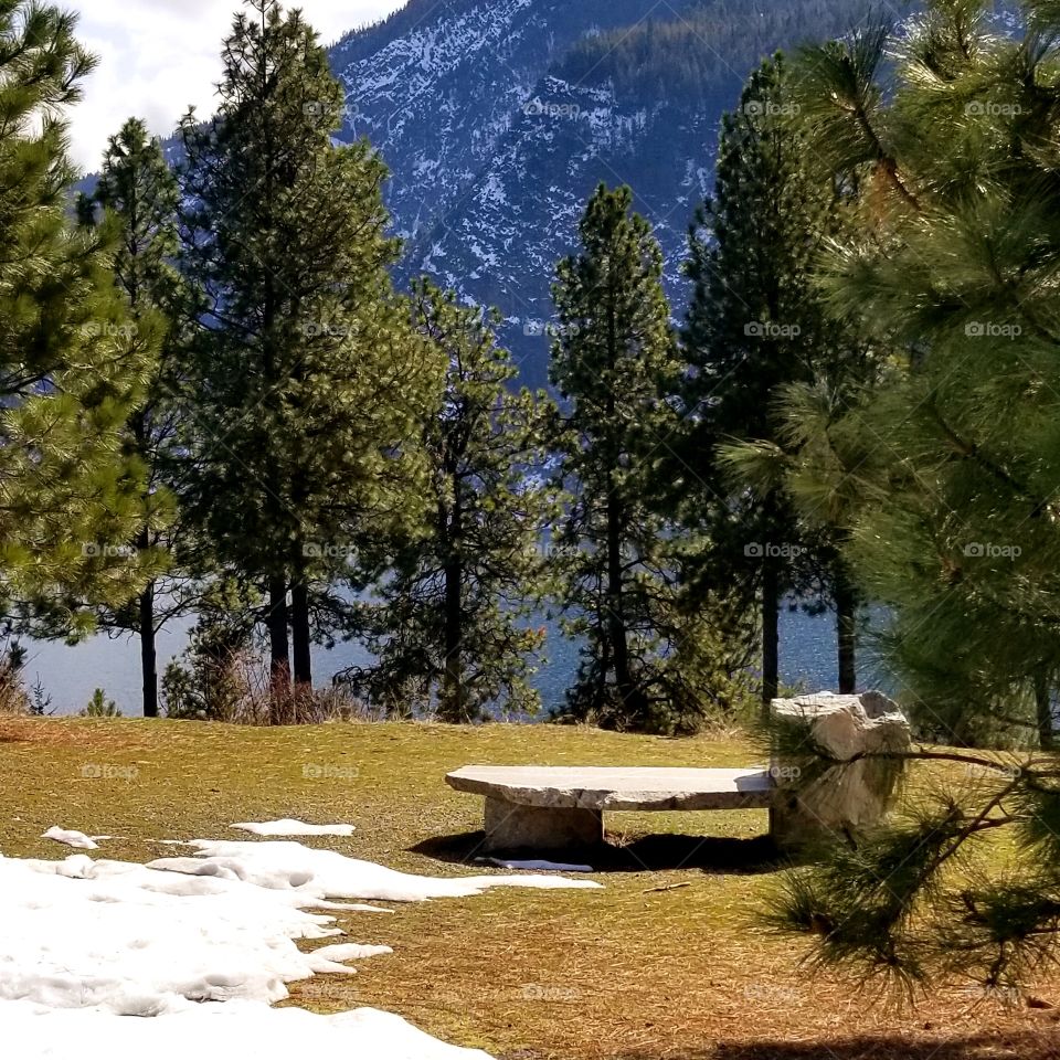 stone bench in a park with snow green trees, lake and snow covered mountain ridge