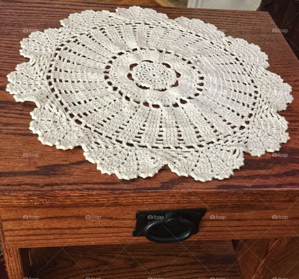 A white tatted doily sitting on a wooden side table. The table has a drawer with a black handle.