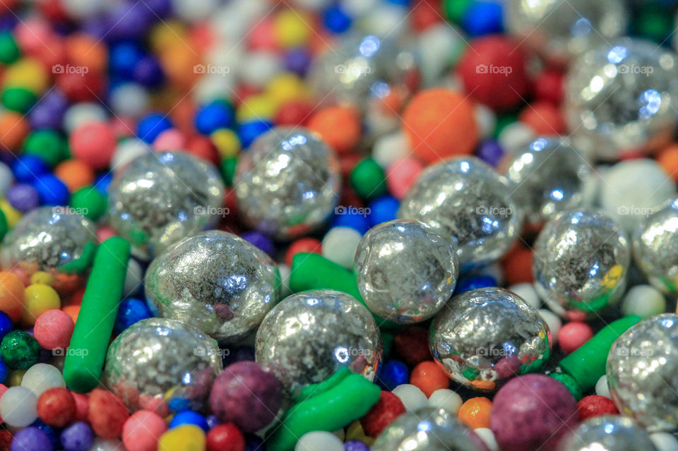 Macro of multi-coloured candy sprinkles of different sizes & shapes. There are also silver balls, dragees, reflecting the other candies & the room.