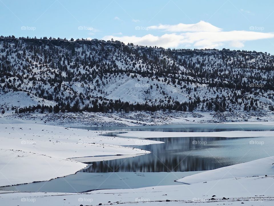 Hills covered in fresh snow and bright skies reflecting on the waters at Juniper Point at Prineville Reservoir in Central Oregon on a winter day.                                                    
