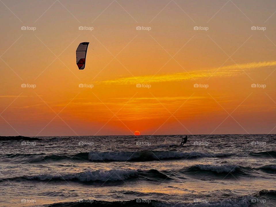 A man riding the waves with a parachute attached to his surf board, with the sunset in the background