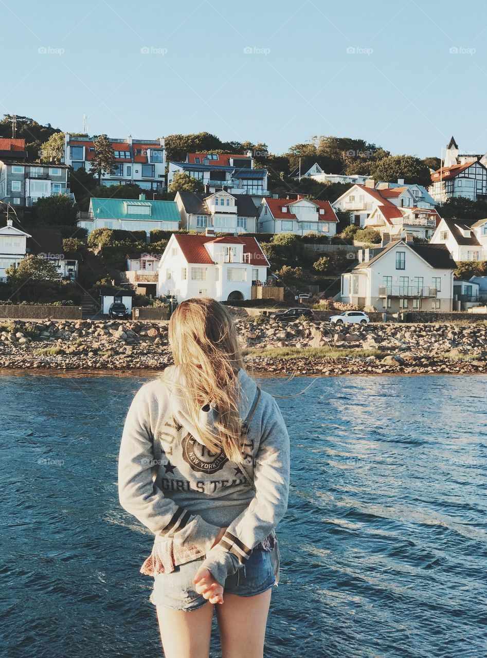 Blonde haired girl standing by the sea looking at the village 