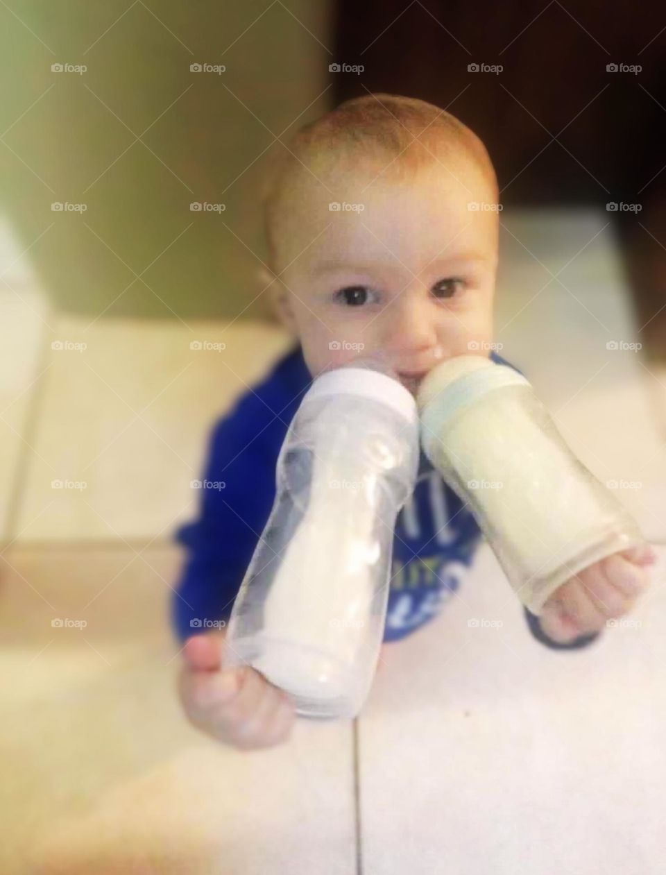 A baby boy walks around drinking from two bottles, holding with both hands. He is wearing a blue shirt. 