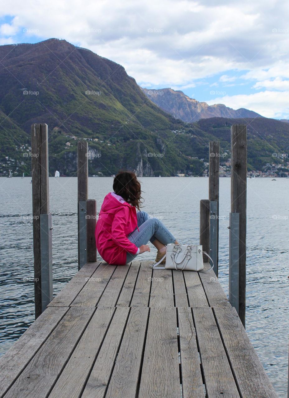 A girl in a pink jacket sits on the dock and enjoys the view of lake Lugano and the surrounding mountain