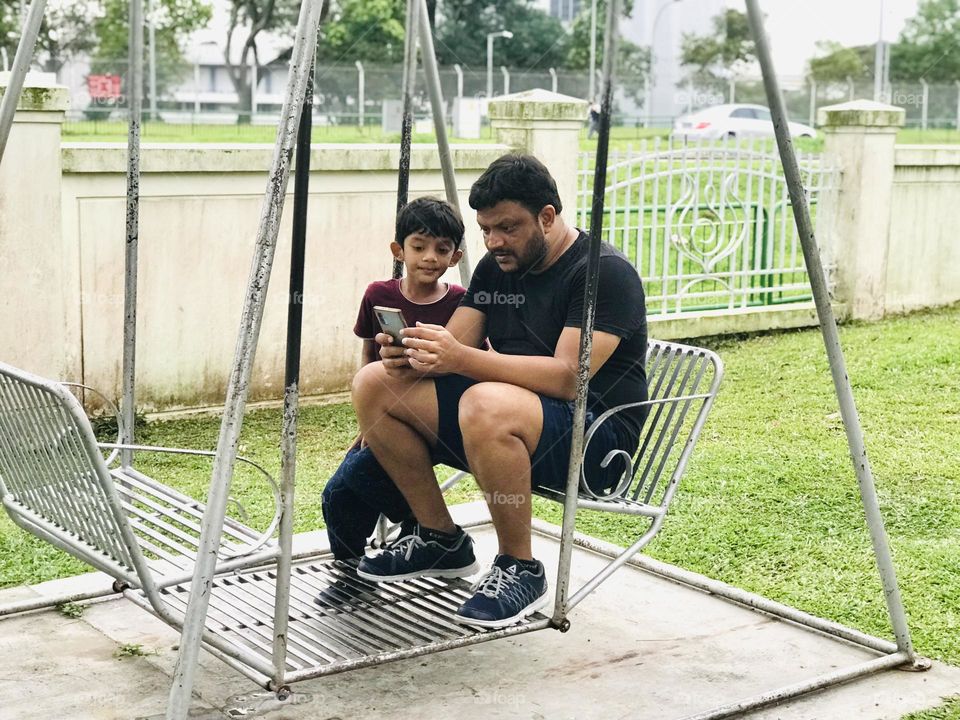 Father and son both sit on a swing and looking into the phone 