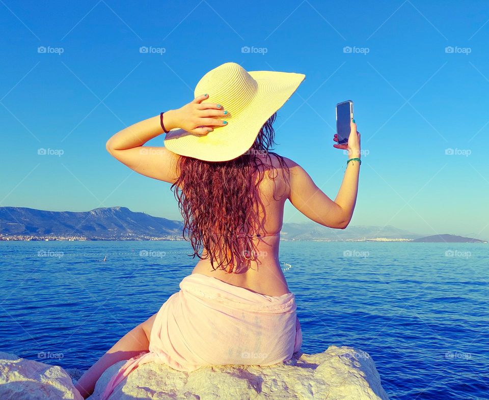 A young woman sits on a rock by the sea and enjoys photographing the seascape with a mobile phone