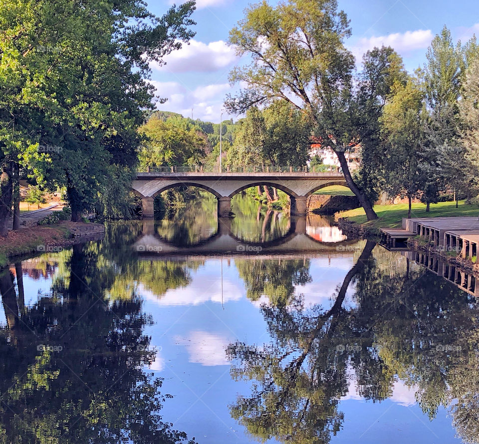A rural scene reflected in the river, including a stone arched bridge, trees, blue sky with fluffy white clouds 