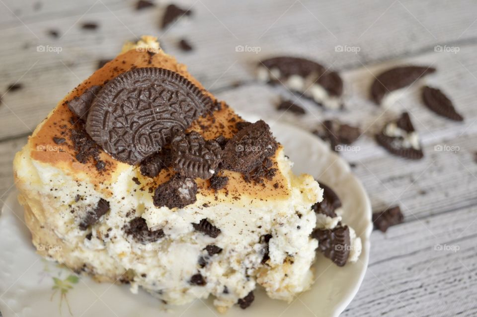 Oreo cookie cheesecake on a white plate with Oreo cookie crumbles on a white rustic background