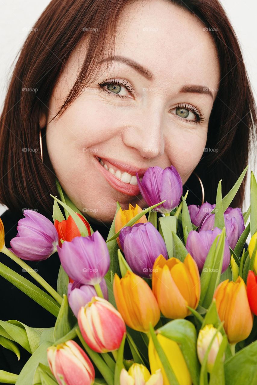 Portrait of a young beautiful brunette who looks into the camera and with a happy smile holds a bouquet of colorful tulips to her face, close-up view from the side.