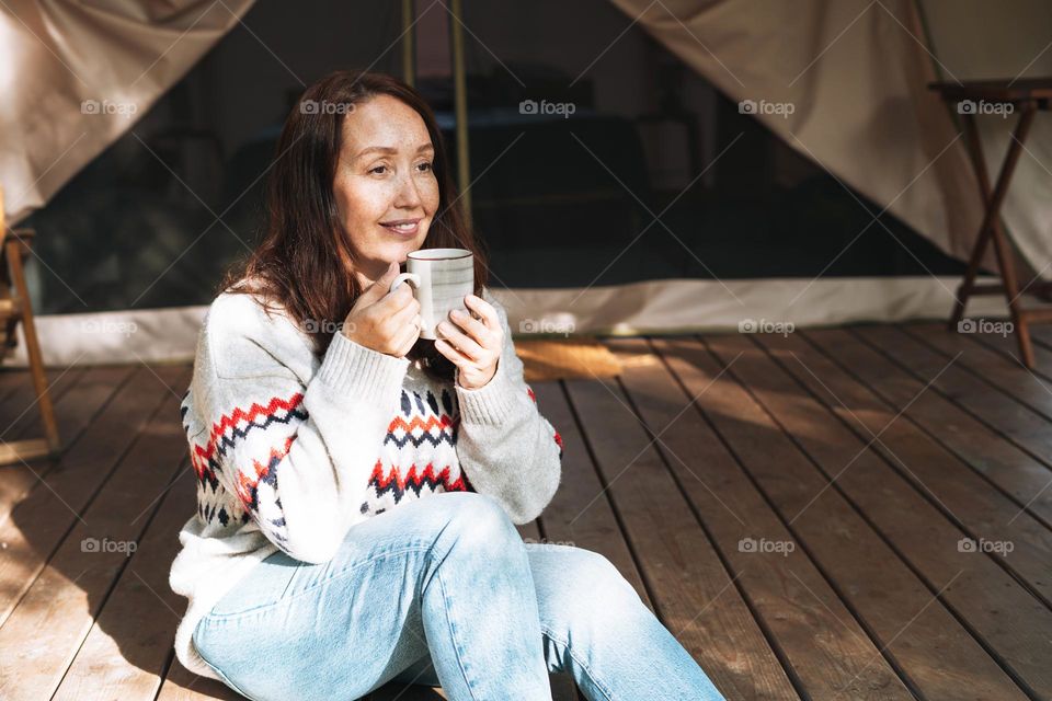 Brunette woman in nordic sweater drinking tea and relaxing in glamping in nature