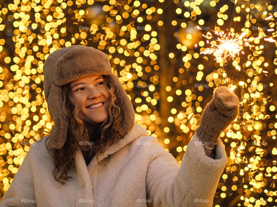 Young beautiful woman with curly hair in fur coat and hat with ear flaps smiling with lights on a background