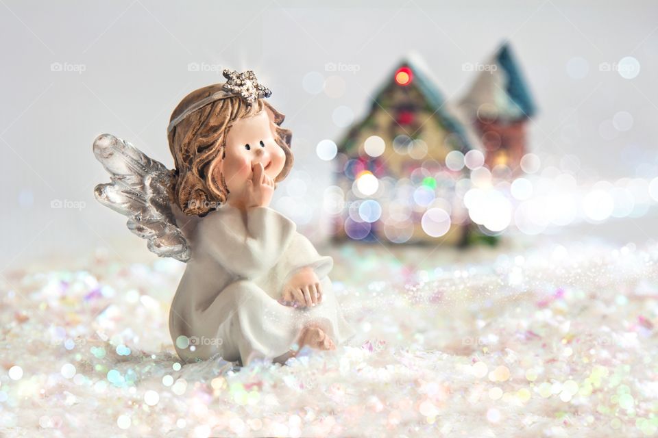 In the foreground, a toy angel sits against a background of artificial snow, and in the background a toy house.