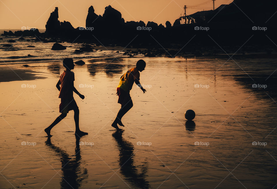 Silhouettes of two boys play soccer on the Indian beach at sunset against the cliffs,  Goa, India