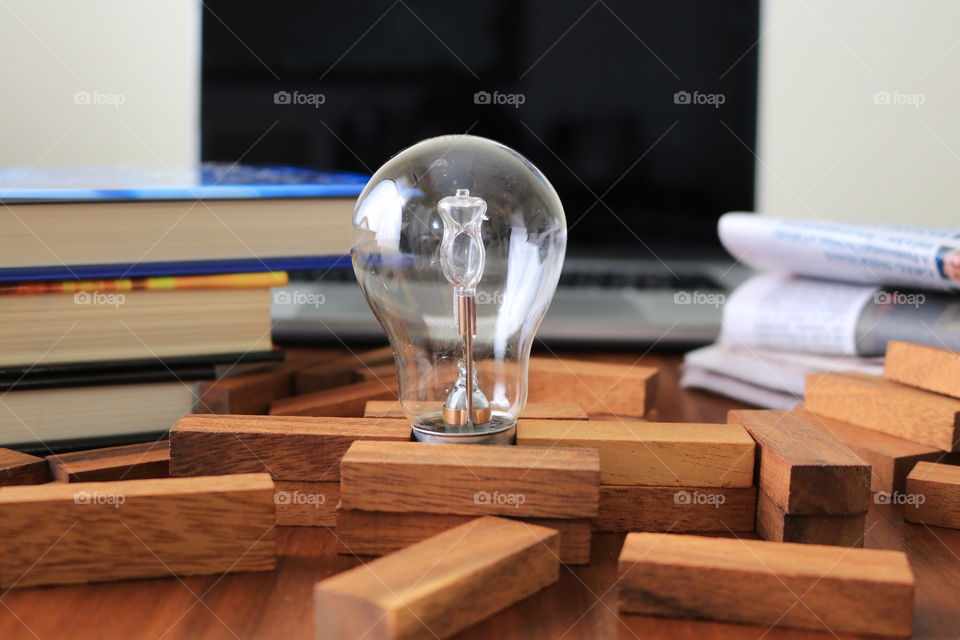 Knowledge, conceptual imagery, light bulb amongst a fallen stack of wood building blocks, with the suggestion of obtaining more knowledge or success by way of the stacked books, newspapers, and computer laptop blurred in background 