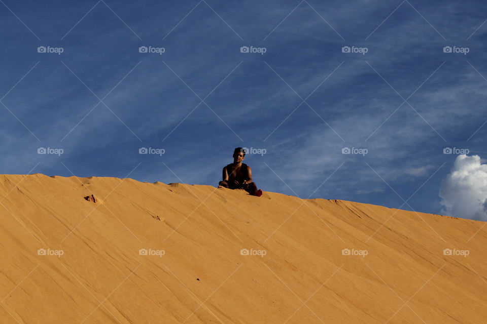 a boy sitting at the top of a sand bank in contrast with vivid blue beautiful sky